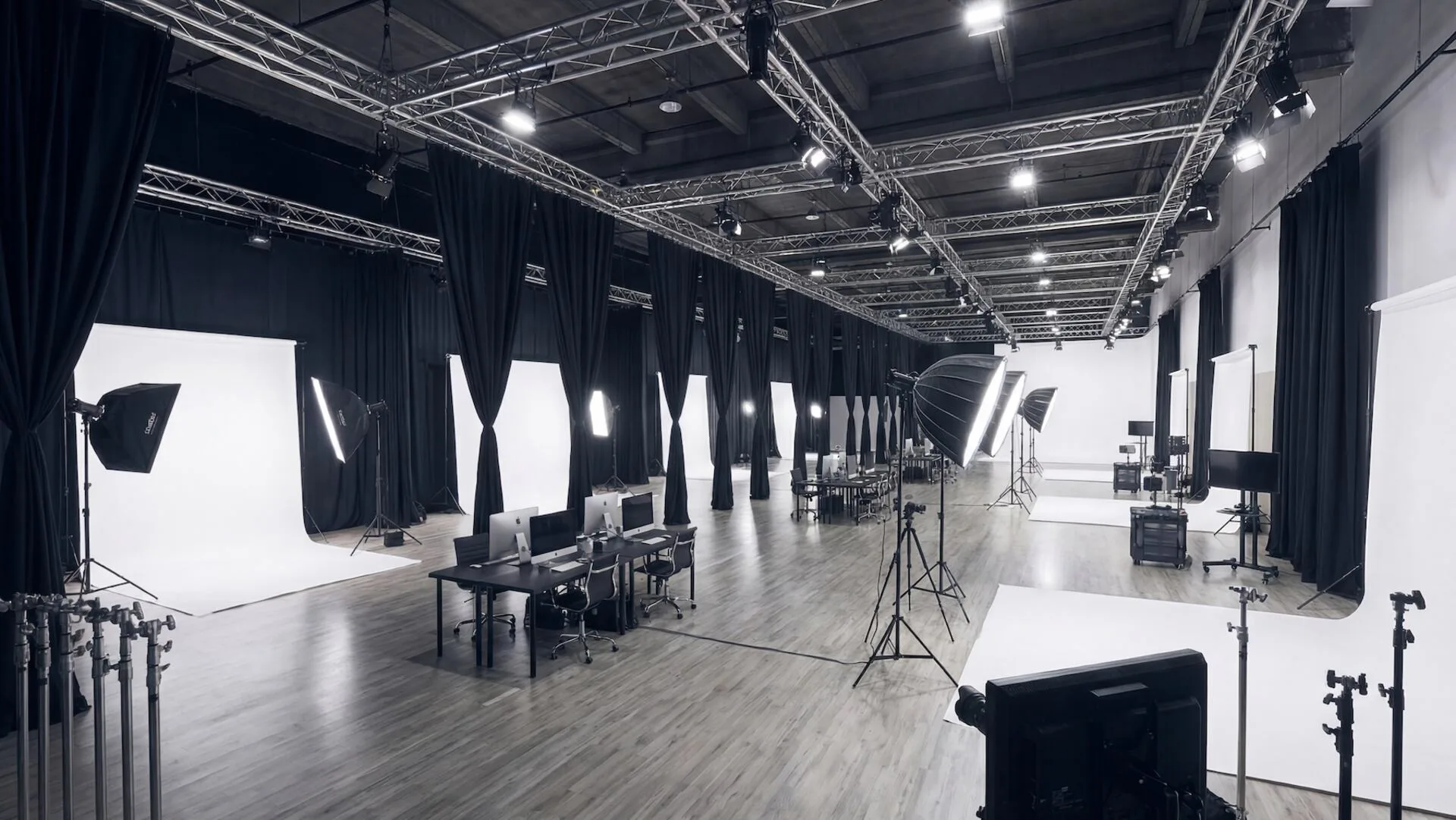A large-scale commercial photography studio featuring a row of modular shooting bays equipped with white seamless backdrops and professional lighting. Each station is paired with a tethered iMac workstation on a polished wood floor, flanked by black floor-to-ceiling privacy curtains and an overhead industrial lighting truss.